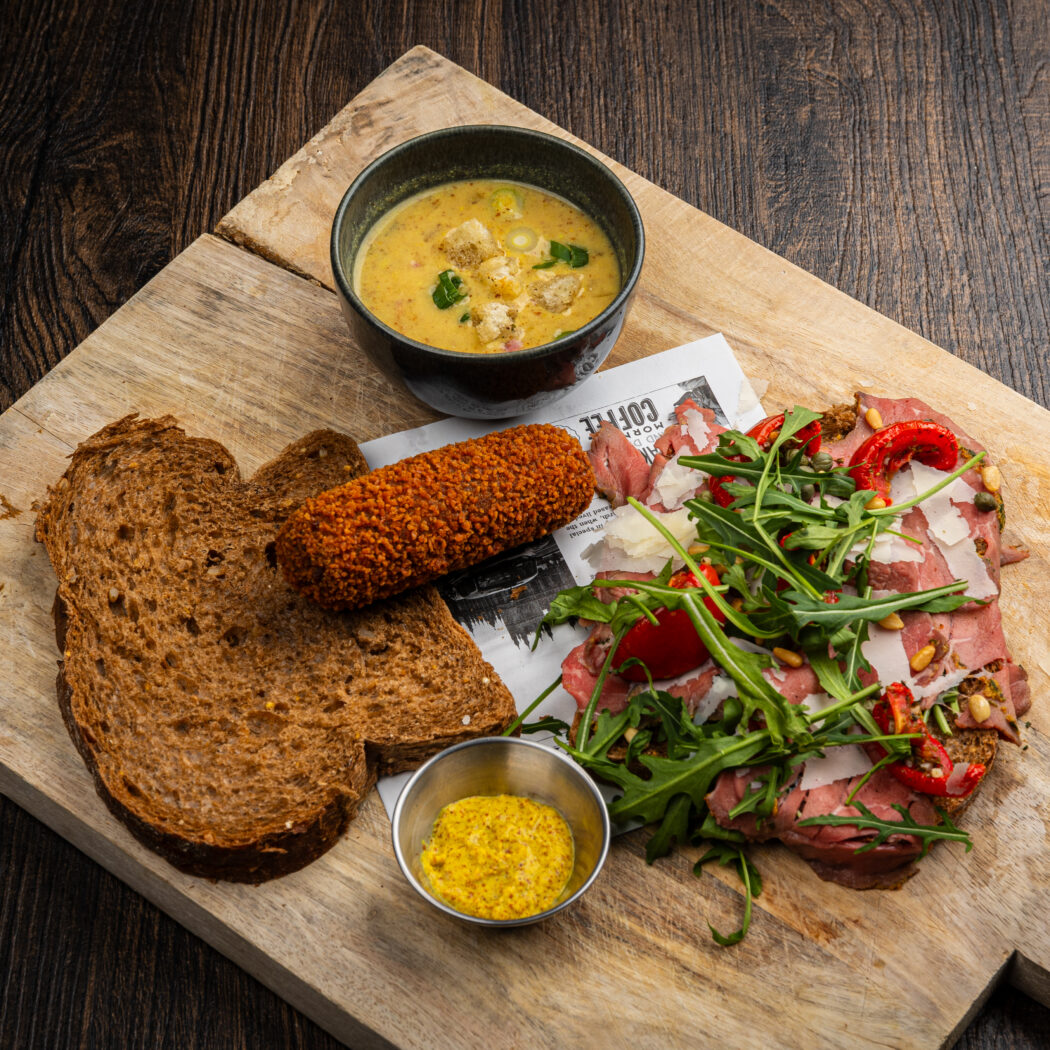 Houten plank met een krokante kroket, donker brood, romige soep en rosbief met rucola en Parmezaan.