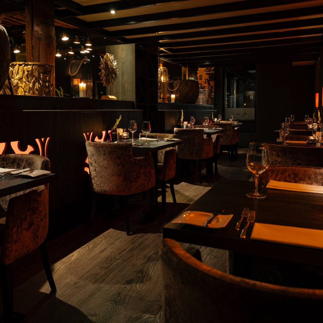 Donker, sfeervol restaurantinterieur met gedekte tafels, fluwelen stoelen en een verlicht decoratief patroon.