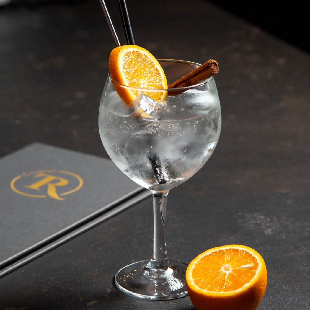 Verfrissende cocktail met sinaasappelschijf, kaneelstokje en rietjes, naast een menukaart met R-logo op een donkere tafel.