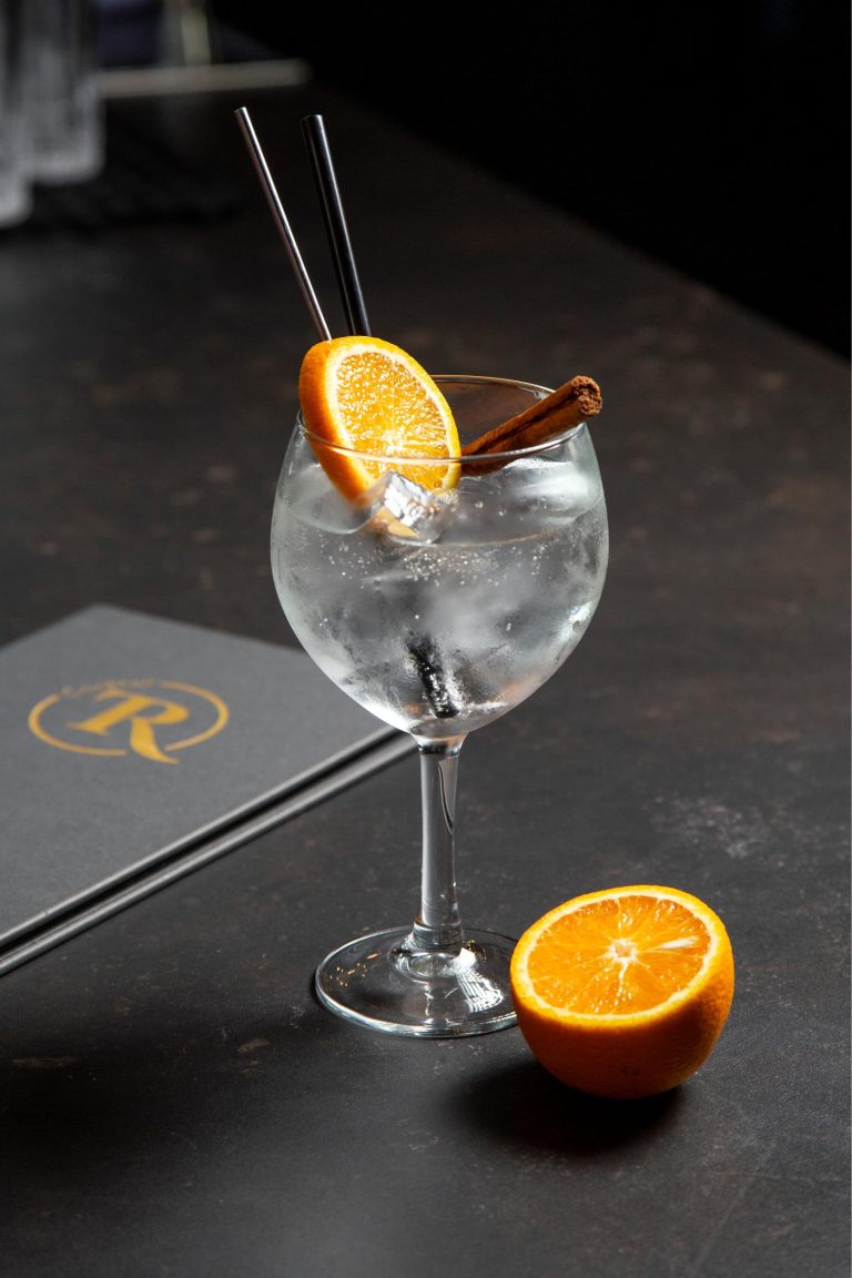 Verfrissende cocktail met sinaasappelschijf, kaneelstokje en rietjes, naast een menukaart met R-logo op een donkere tafel.