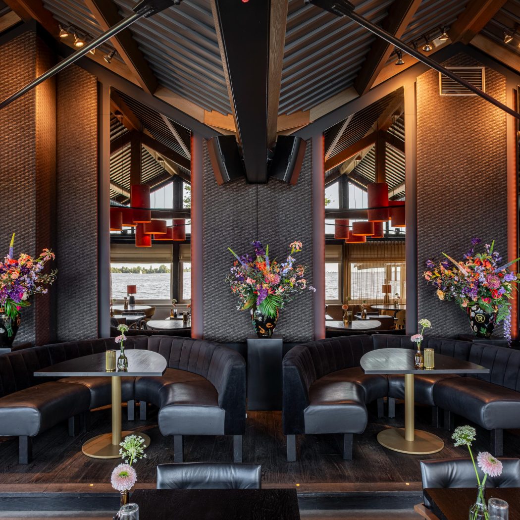 Modern restaurant met donkere, gebogen zitbanken, ronde tafels en kleurrijke bloemstukken, met waterzicht.