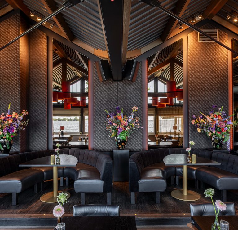 Modern restaurant met donkere, gebogen zitbanken, ronde tafels en kleurrijke bloemstukken, met waterzicht.