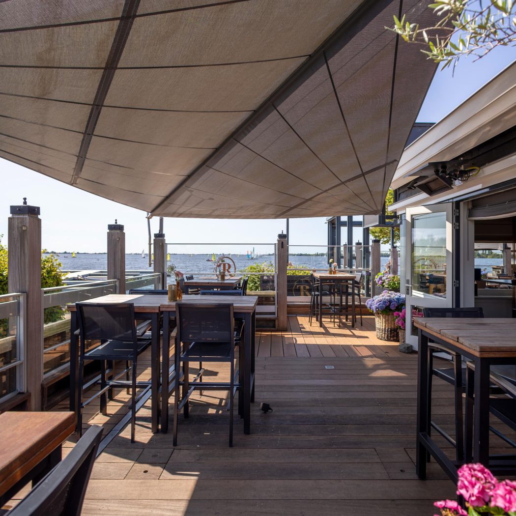 Zonnig buitenterras van een restaurant met houten vlonder, tafels en een groot zonnescherm, met uitzicht op het water en zeilboten.