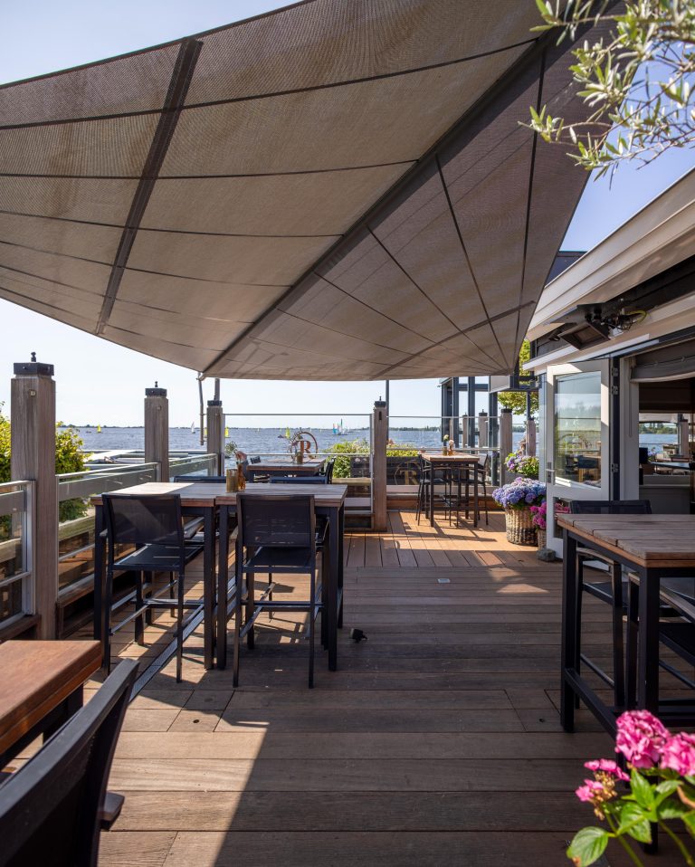 Zonnig buitenterras van een restaurant met houten vlonder, tafels en een groot zonnescherm, met uitzicht op het water en zeilboten.