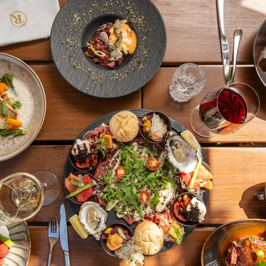 Luxe maaltijd met diverse gerechten: vis, vlees, oesters, carpaccio, salade, wijnen en een servet met R logo op een houten tafel.