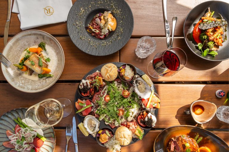 Luxe maaltijd met diverse gerechten: vis, vlees, oesters, carpaccio, salade, wijnen en een servet met R logo op een houten tafel.