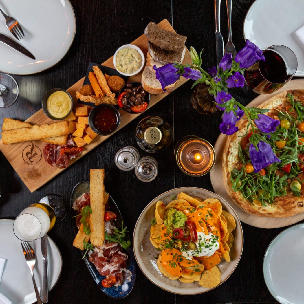 Gezellige donkere tafel vol met verse oesters, charcuterie, flammkuchen, nachos, wijn en paarse bloemen.