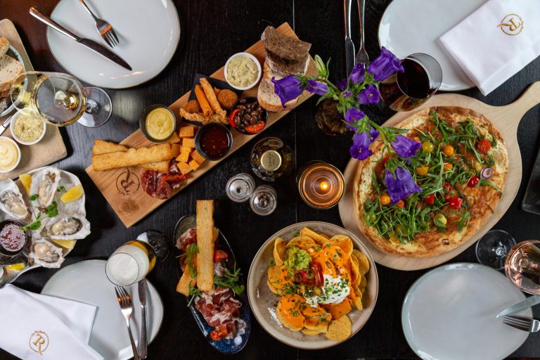 Gezellige donkere tafel vol met verse oesters, charcuterie, flammkuchen, nachos, wijn en paarse bloemen.