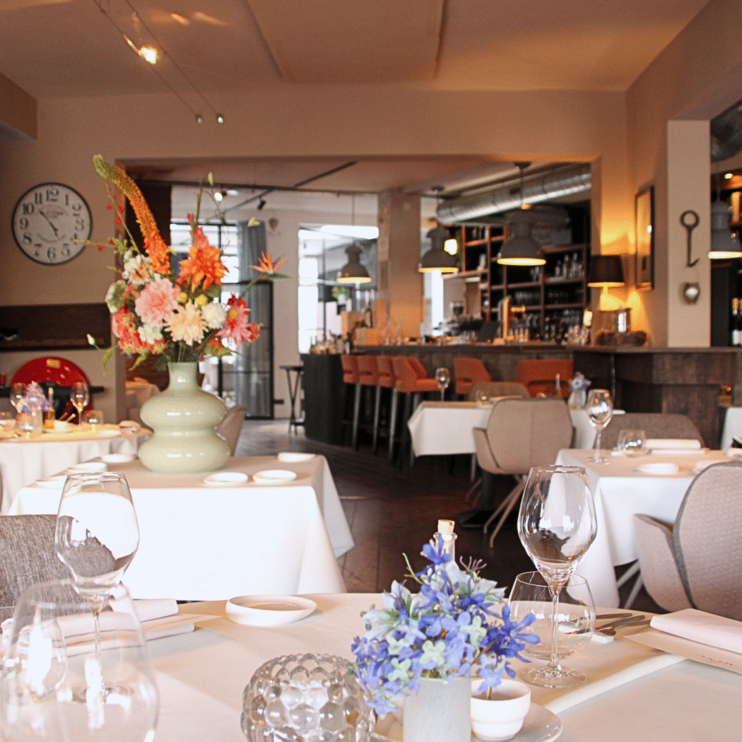 Sfeervol restaurant met gedekte tafels, kleurrijke bloemstukken en een bar met oranje stoelen.