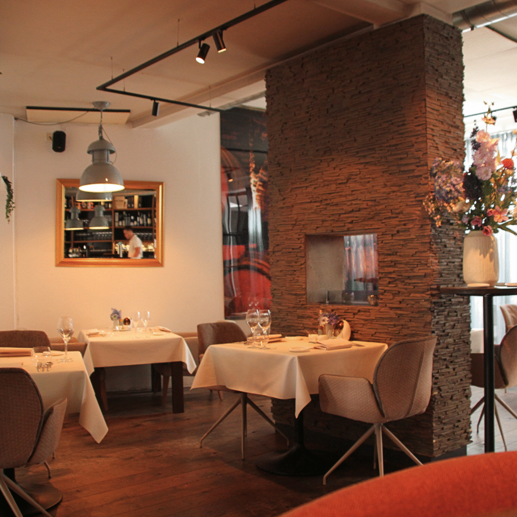 Restaurant interieur met tafels met wit linnen, een stenen pilaar met bloemen en een spiegel die een bar reflecteert.