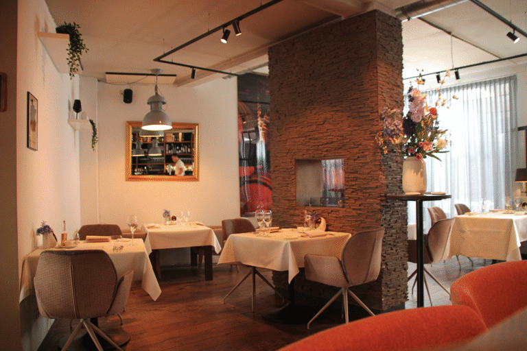 Restaurant interieur met tafels met wit linnen, een stenen pilaar met bloemen en een spiegel die een bar reflecteert.