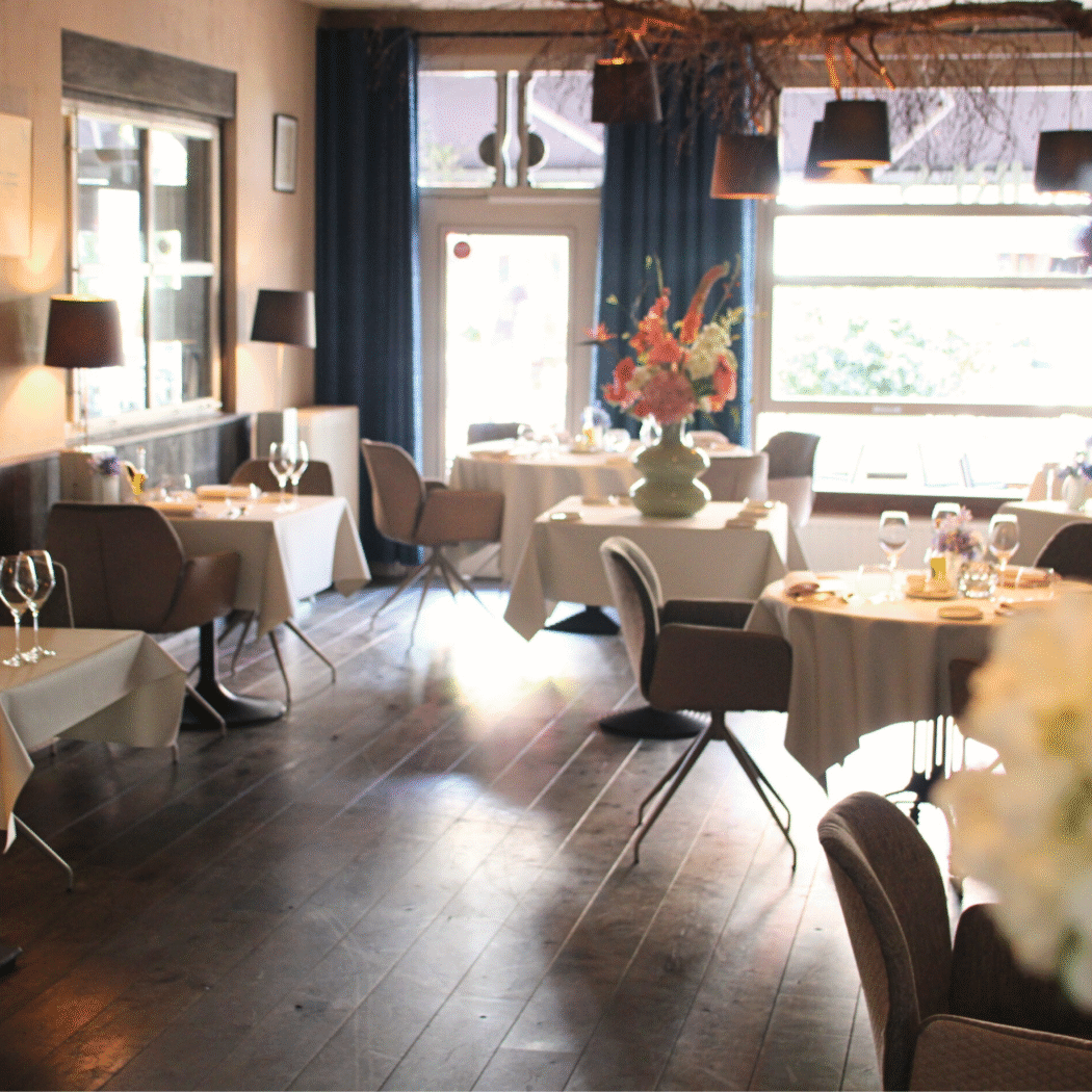Sfeervol restaurant interieur met gedekte tafels, bruine stoelen, bloemstukken en veel daglicht.