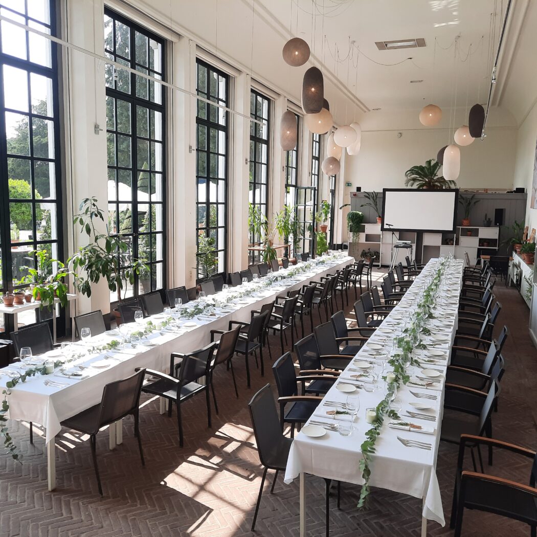 Lichte evenementenzaal met twee lange gedekte tafels versierd met groene slingers, grote ramen en projectiescherm.