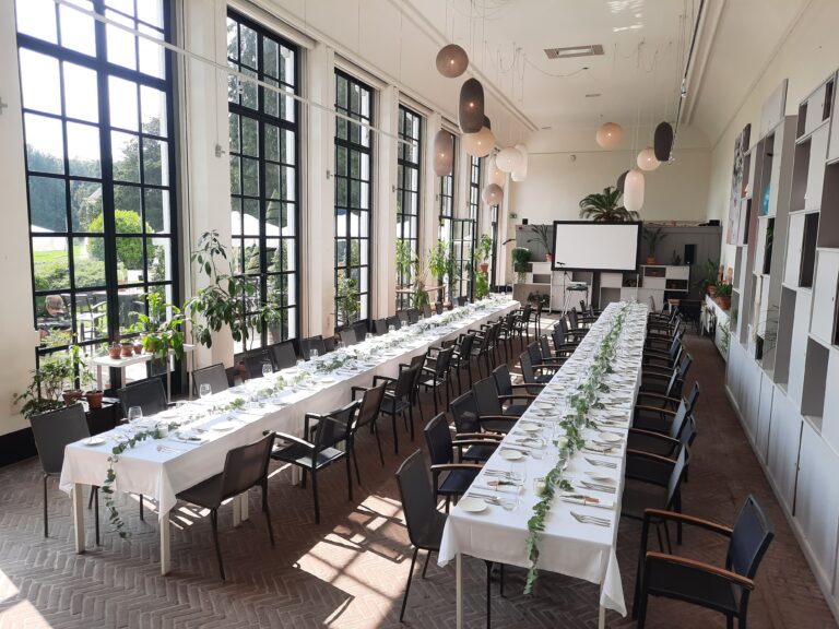 Lichte evenementenzaal met twee lange gedekte tafels versierd met groene slingers, grote ramen en projectiescherm.