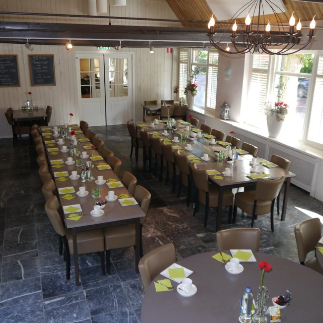 Ruime evenementenzaal met lange en ronde gedekte tafels, een kroonluchter en ramen met witte jaloezieën.