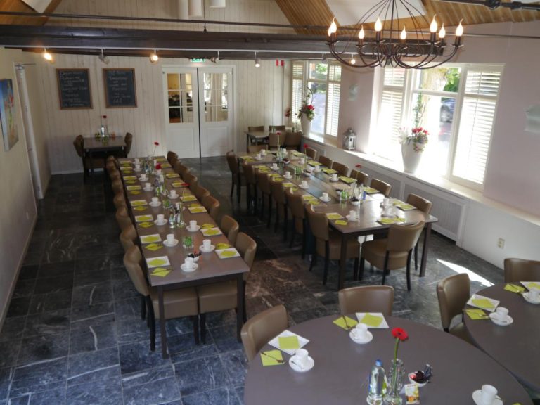 Ruime evenementenzaal met lange en ronde gedekte tafels, een kroonluchter en ramen met witte jaloezieën.