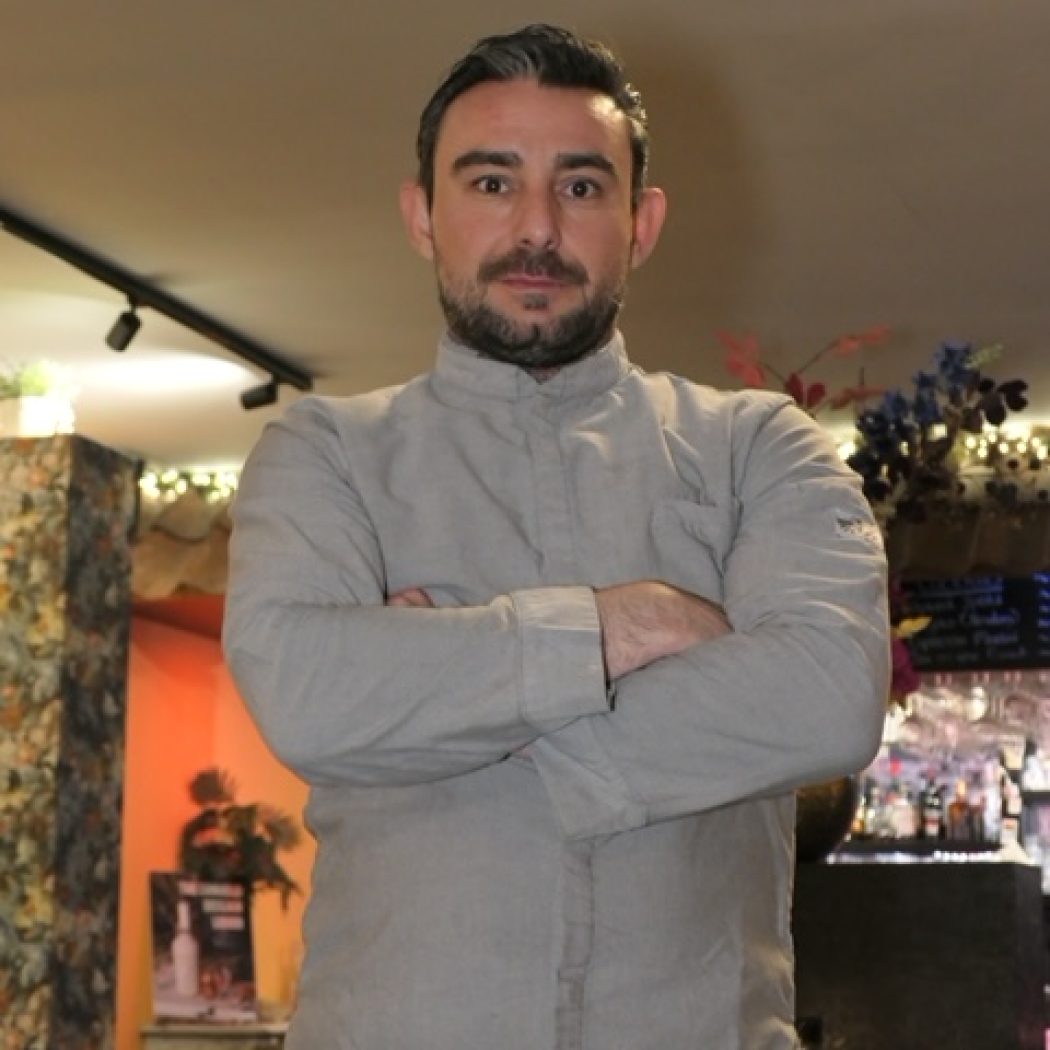 Restaurateur met baard en grijs werkoverhemd, met gekruiste armen in een warme ambiance.