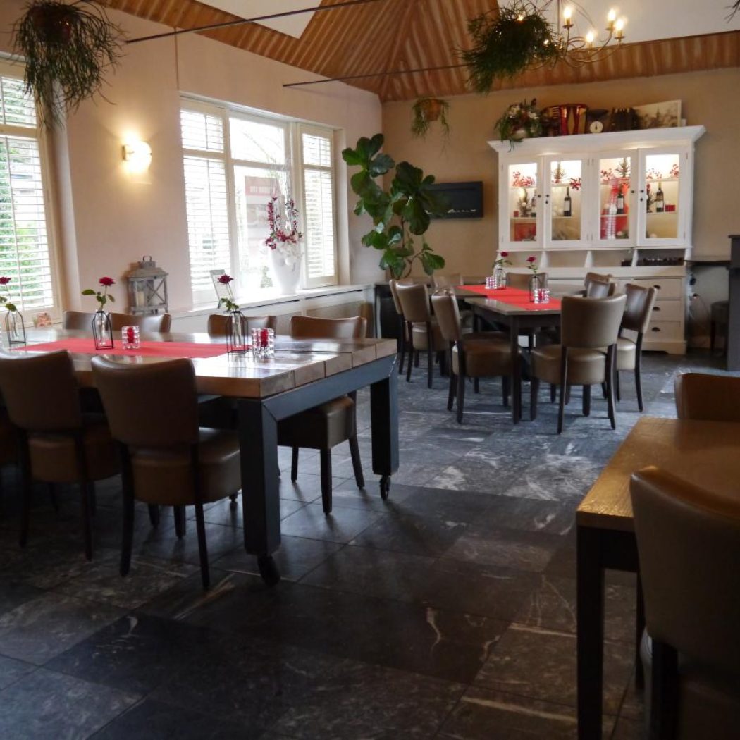 Sfeervol restaurantinterieur met lange houten eettafel, bruine stoelen, rode tafeldecoratie en een verlichte buffetkast.