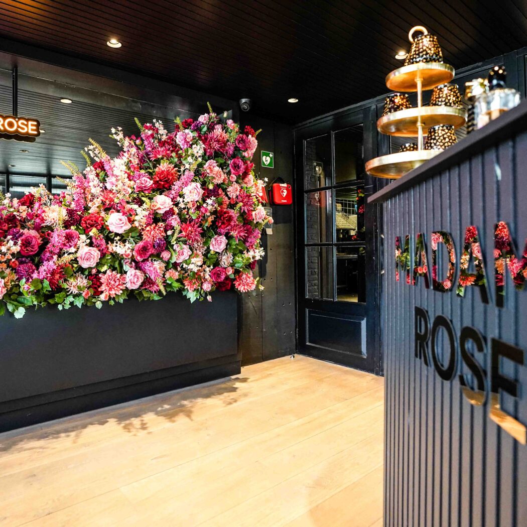 Weelderige roze bloemenwand met neon XOXO #MADAMEROSE en een toonbank met MADAME ROSE logo.