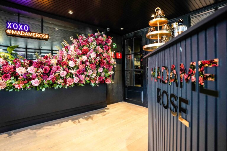 Weelderige roze bloemenwand met neon XOXO #MADAMEROSE en een toonbank met MADAME ROSE logo.