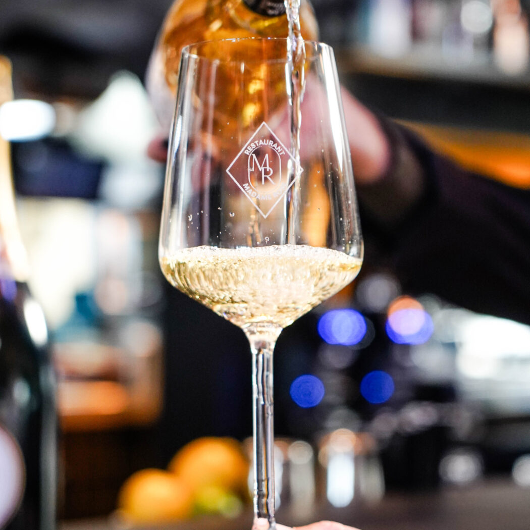 Hand schenkt mousserende wijn in een wijnglas met het logo “RESTAURANT MADAME R”.