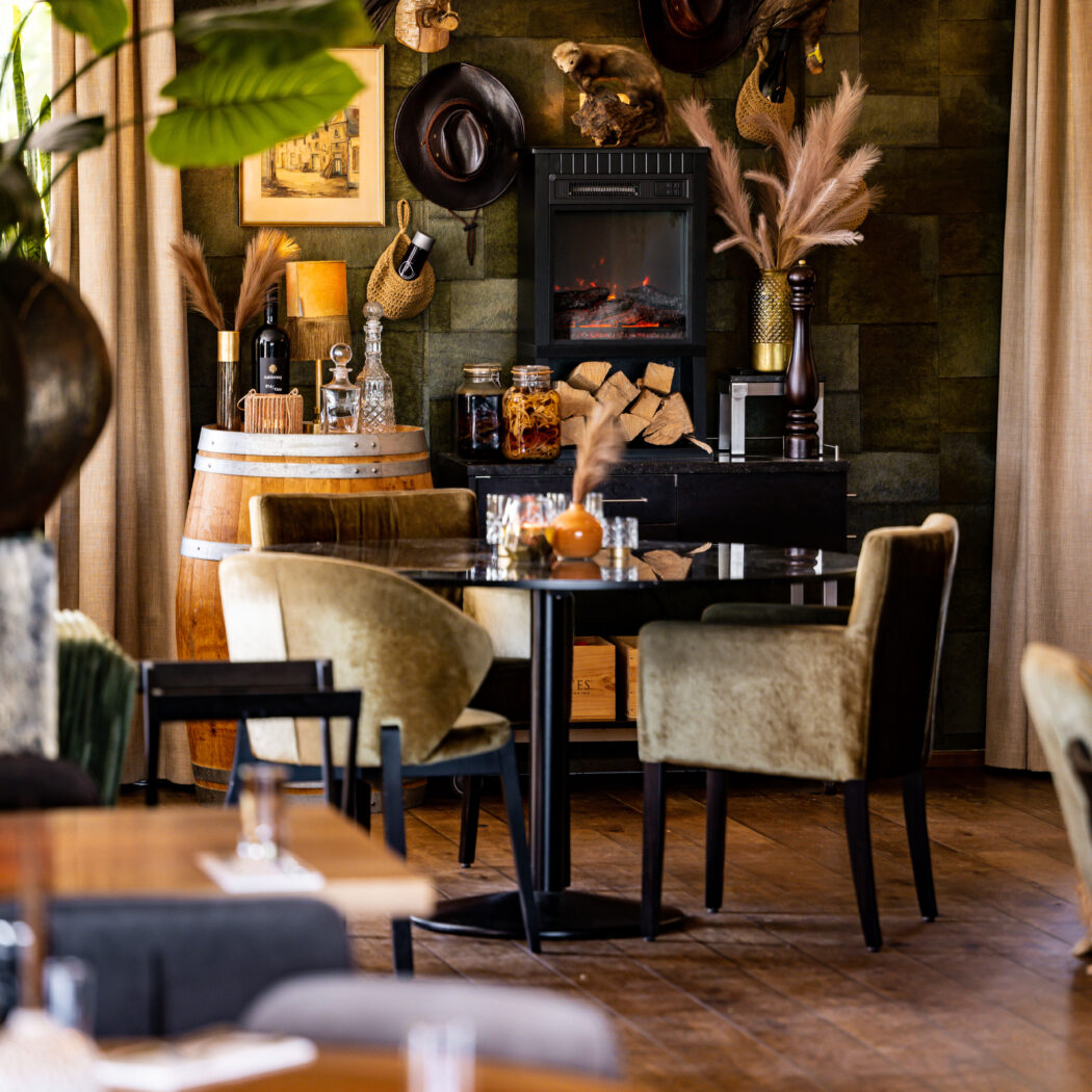 Groen interieur met western-decoratie, brandende haard, houten vat en fluwelen stoelen aan tafels.