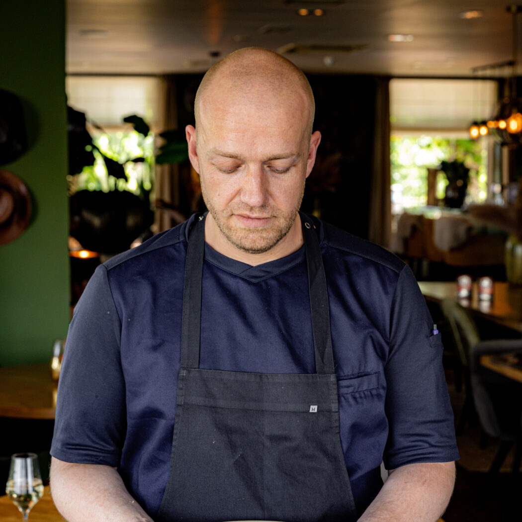 Chef in blauw uniform en zwart schort toont zorgvuldig opgemaakt gerecht op beige bord.