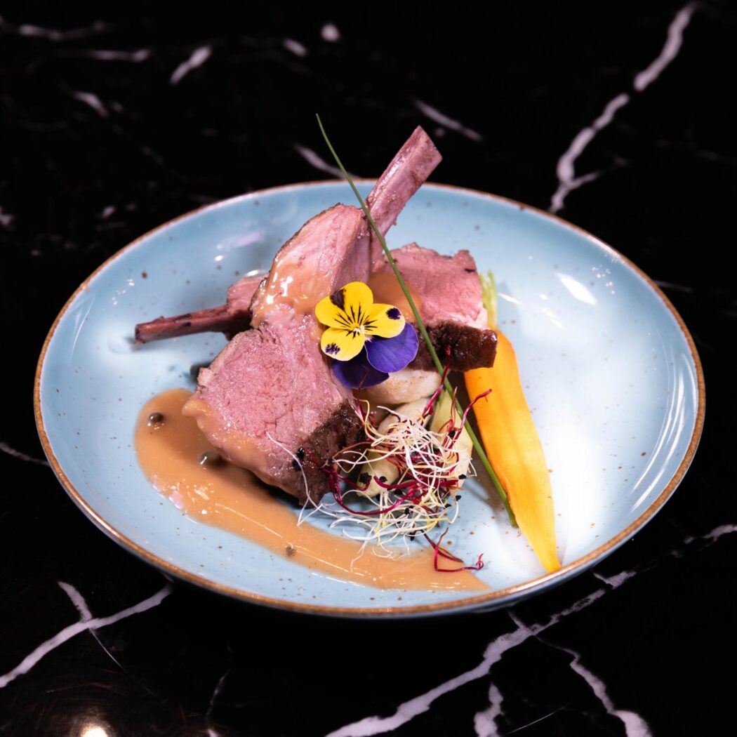 Mooi opgemaakt lamsrack met saus, wortel en paars-gele eetbare bloemen op blauw bord.