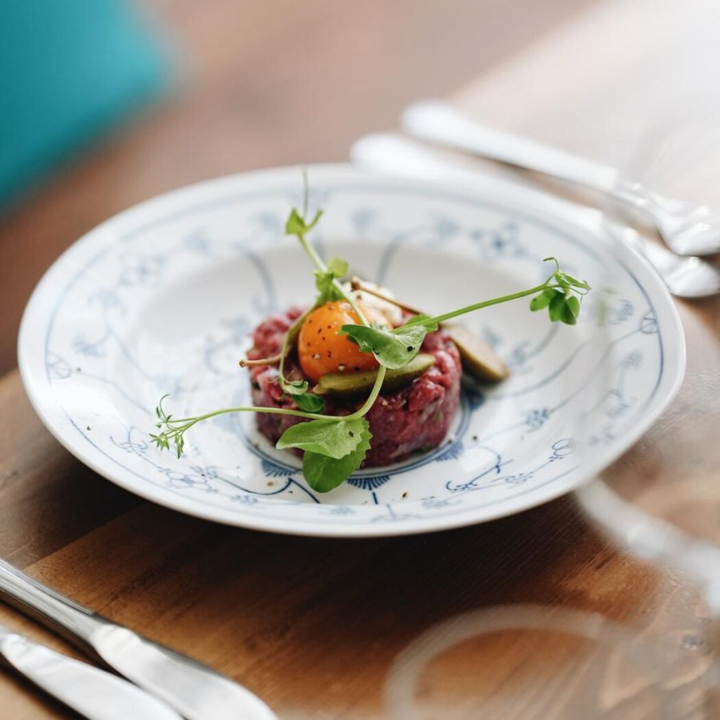 Steak tartaar met rauwe eidooier, groene kiemen en augurken, geserveerd op een wit-blauw porseleinen bord op een houten tafel.