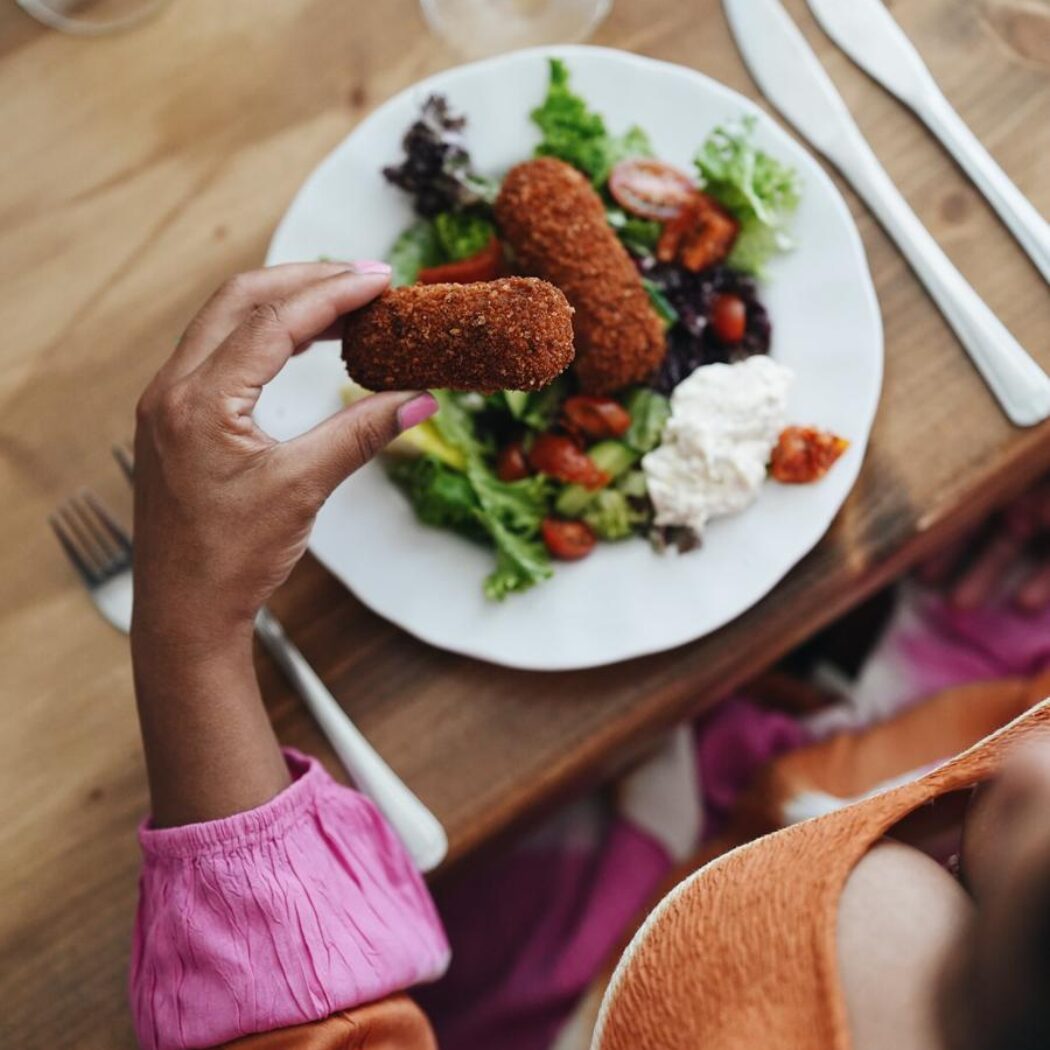 Een hand met roze nagellak houdt een krokante kroket boven een bord met salade op een houten tafel.