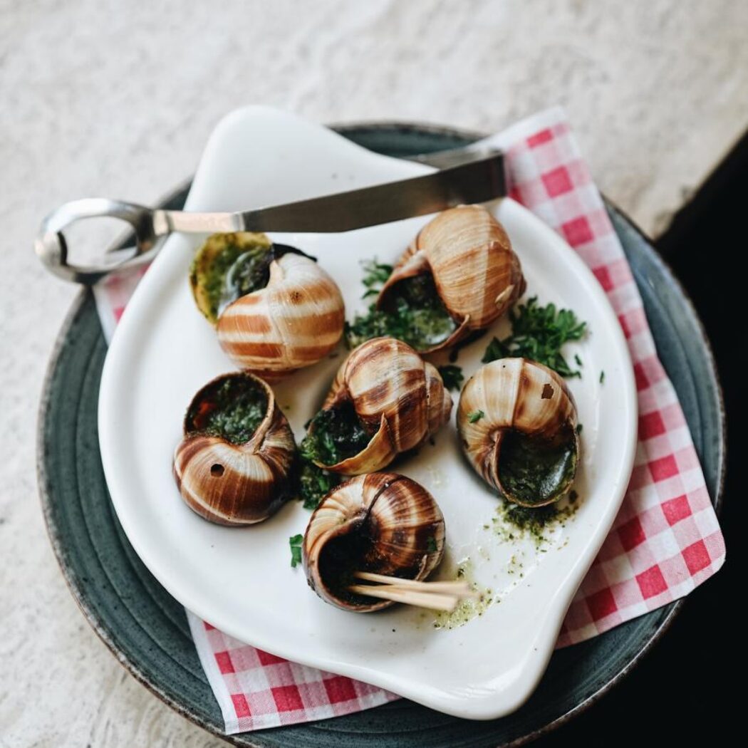 Zes escargots met kruidenboter in schelpen, geserveerd op een wit bord met zilveren tang en rood-wit geruit servet.