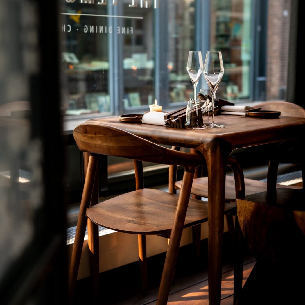Houten restauranttafel gedekt voor twee met champagneglazen en een brandende kaars, bij raam met Fine Dining reflectie.