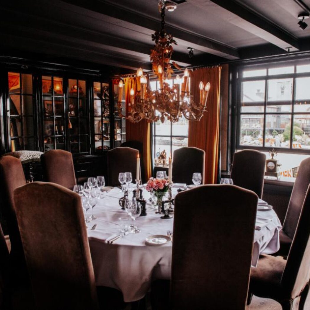 Sfeervolle, donkere eetzaal met gedekte ronde tafel, bruine stoelen, roze bloemen en een antieke kroonluchter.