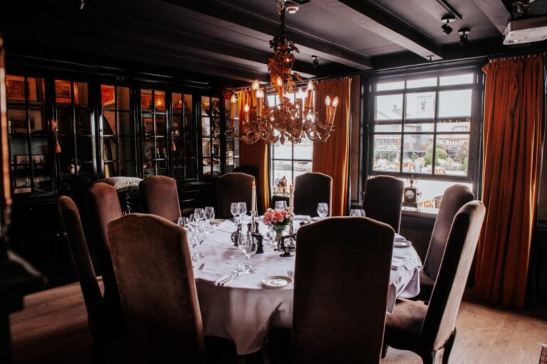 Sfeervolle, donkere eetzaal met gedekte ronde tafel, bruine stoelen, roze bloemen en een antieke kroonluchter.