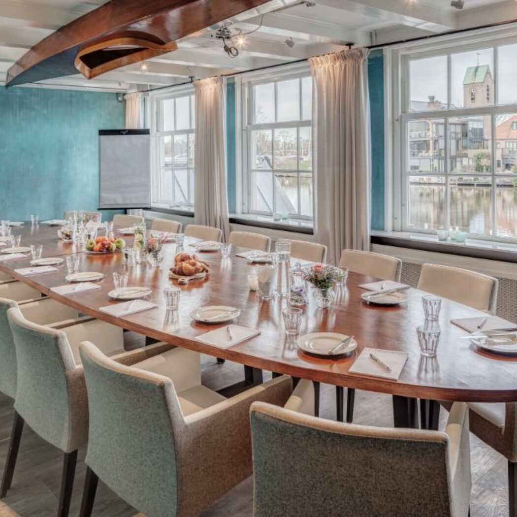 Sfeervolle vergaderzaal met lange houten tafel, klaargezet voor bijeenkomst, grachtzicht en decoratieve boot aan het plafond.