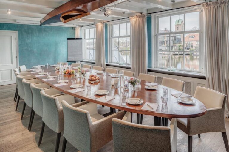 Sfeervolle vergaderzaal met lange houten tafel, klaargezet voor bijeenkomst, grachtzicht en decoratieve boot aan het plafond.