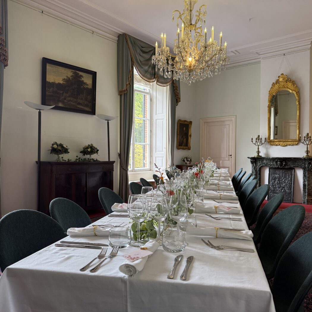 Lange, formeel gedekte eettafel in een statige eetzaal met kroonluchter, open haard en grote ramen.