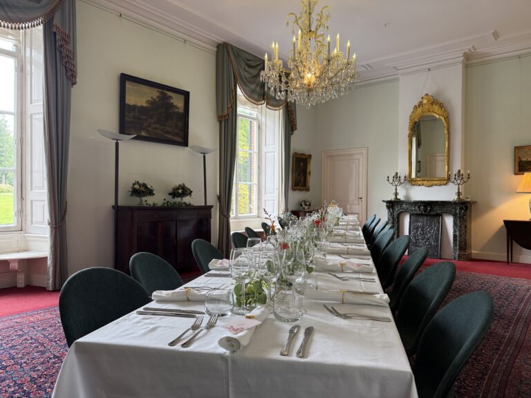 Lange, formeel gedekte eettafel in een statige eetzaal met kroonluchter, open haard en grote ramen.