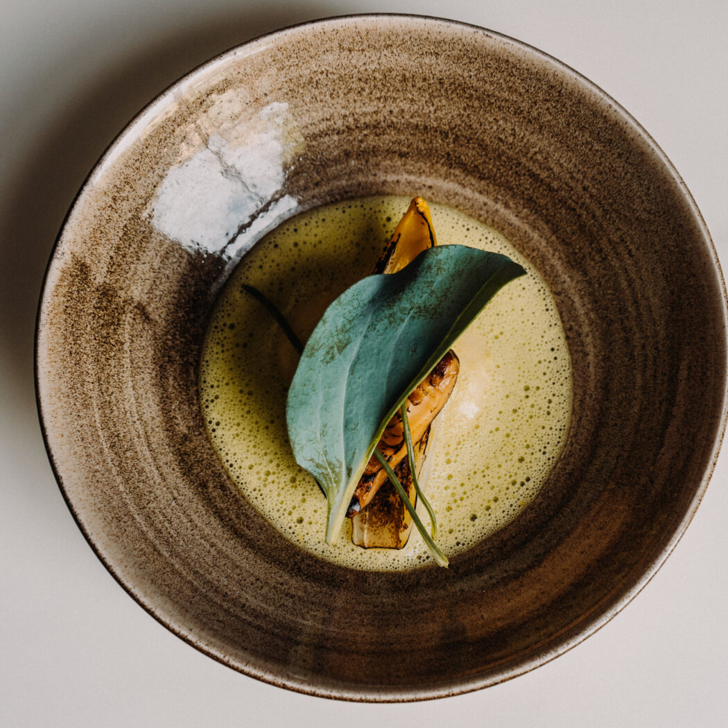 Culinair gerecht in een gespikkelde aardewerken kom met groene schuimsaus, geroosterde groenten en een groot groen blad.