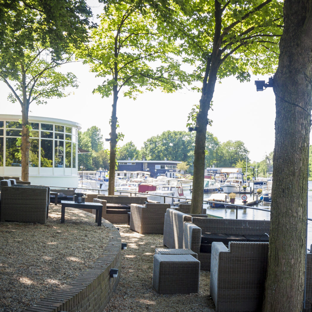Schaduwrijk terras aan het water met loungestoelen en een gebouw met grote ramen, langs een kanaal met boten.