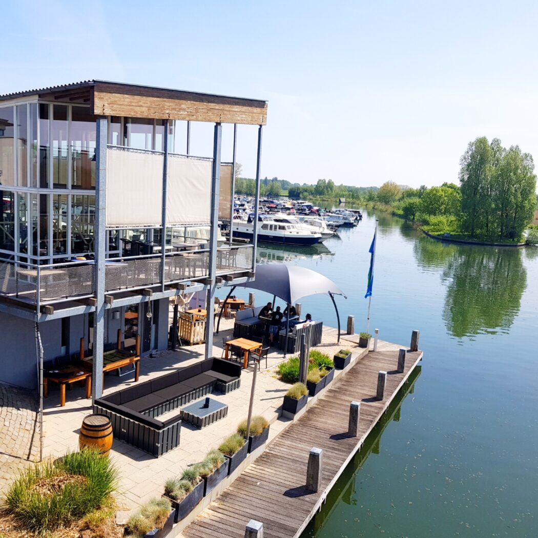Modern glazen restaurant met houten accenten, terras met loungehoeken aan een jachthaven.
