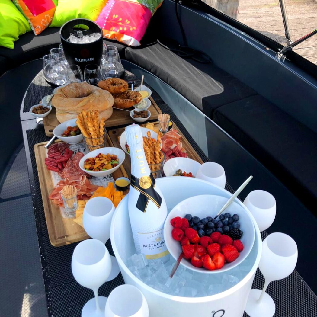 Luxe picknick op bootdek met een charcuterieplank, Moët & Chandon champagne, bessen en felle kussens.