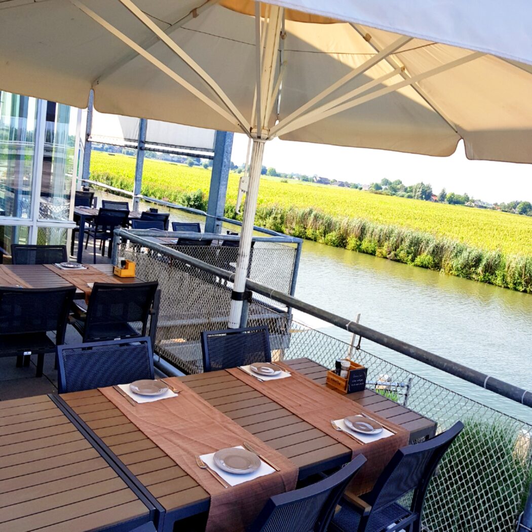 Zonnig restaurantterras met gedekte tafels en parasol, uitzicht op een kanaal en groene velden.