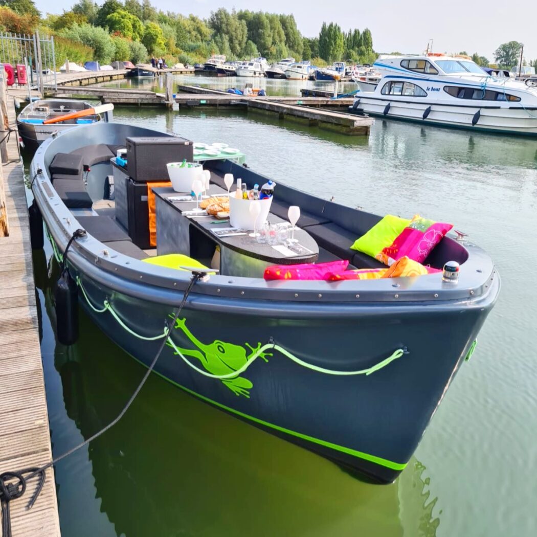 Donkergrijze sloep met groene kikkeropdruk en een picknicktafel vol champagneglazen, aangemeerd aan een houten steiger.