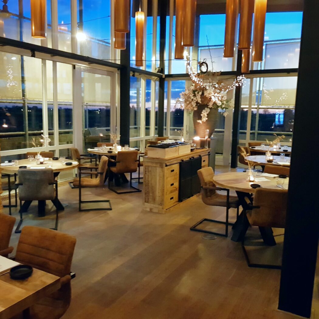Sfeervol restaurant met ronde houten tafels, bruine stoelen, opvallende lampen en grote ramen bij zonsondergang.