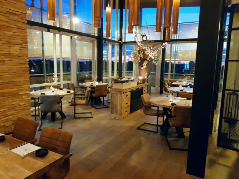 Sfeervol restaurant met ronde houten tafels, bruine stoelen, opvallende lampen en grote ramen bij zonsondergang.