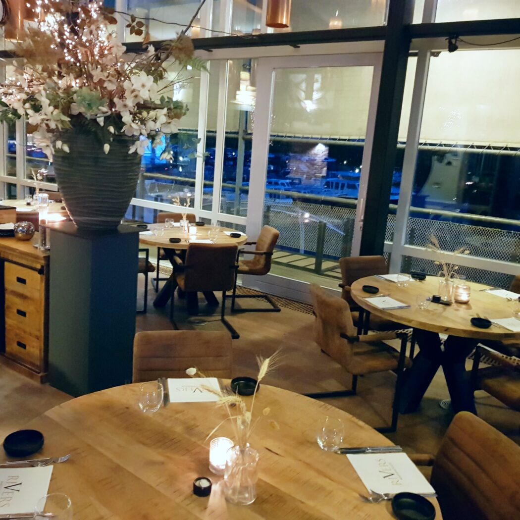Sfeervol restaurant met houten tafels, bruine stoelen, grote ramen, bloemstuk met lichtjes en RIVERS menus.