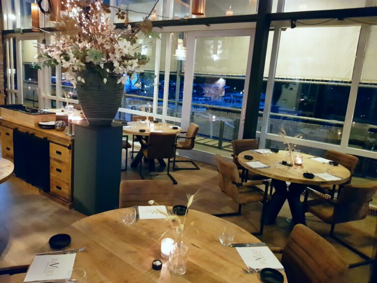 Sfeervol restaurant met houten tafels, bruine stoelen, grote ramen, bloemstuk met lichtjes en RIVERS menus.