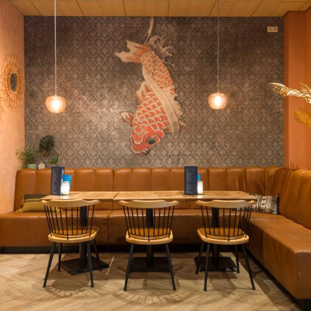 Modern restaurant met cognackleurige lederen zitbank, houten tafels en stoelen, en grote oranje koi karper op de patroonwand.
