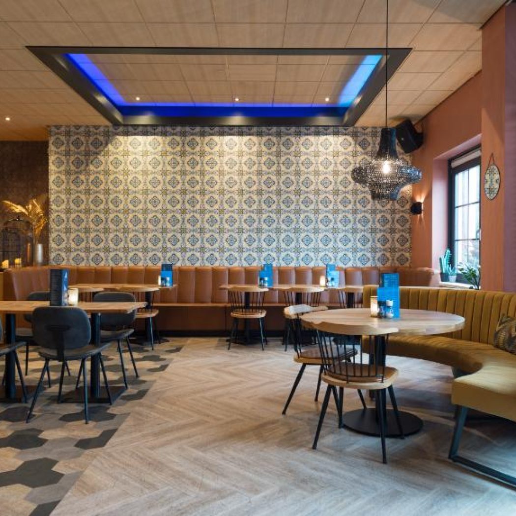 Sfeervol restaurant met bruine en gele banken, houten tafels, een tegelwand met patroon en blauwe plafondverlichting.